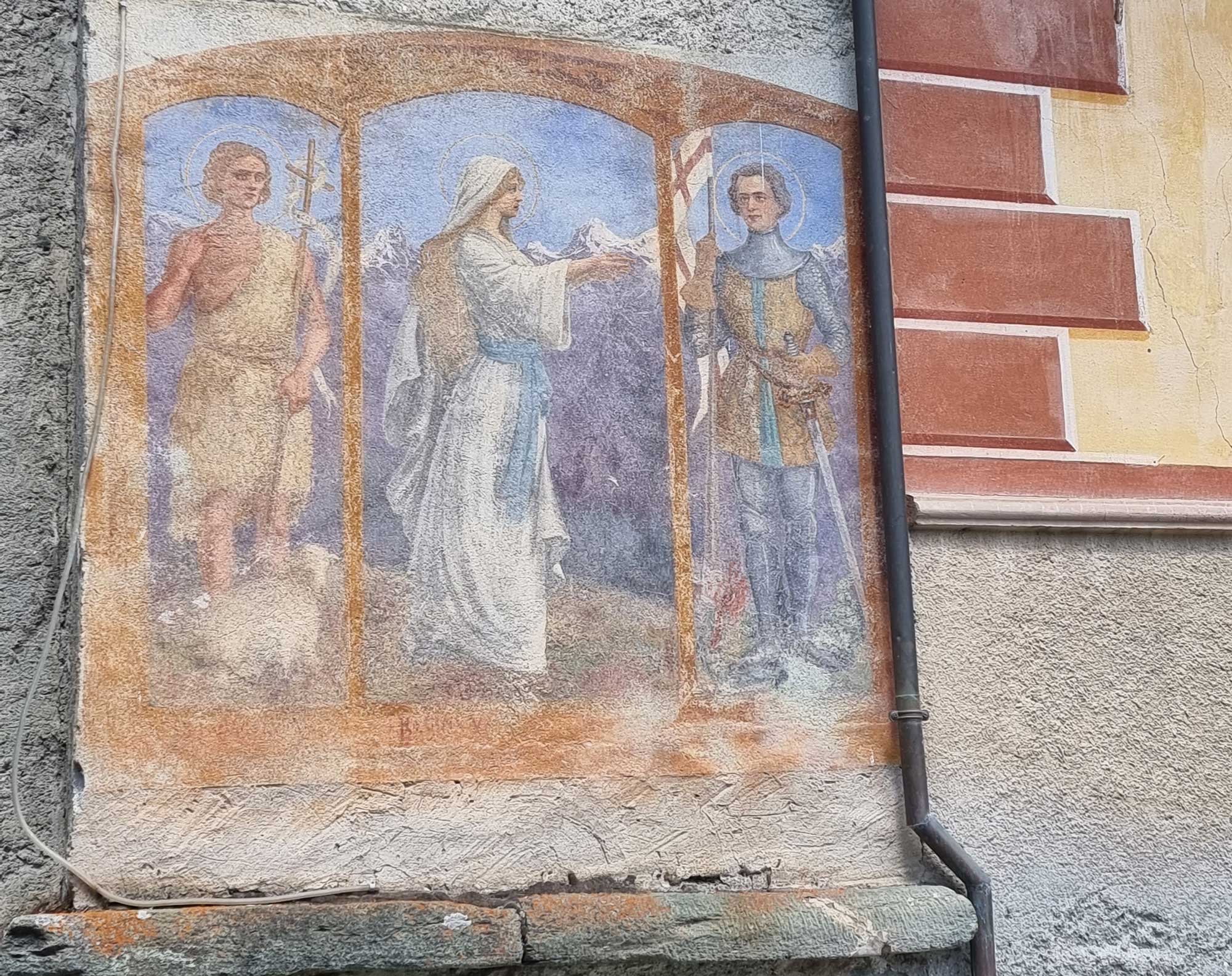 Composizione a trittico raffigurante al centro la Vergine REGINA VALLIS, vestita di bianco e cinta in vita da una fascia celeste. A sinistra è San Giovanni Battista con i classici attributi iconografici (abito di pelo di cammello, croce con cartiglio ECCE AGNVS DEI) e agnello ai suoi piedi. A destra è San Giorgio nell’atto di sconfiggere con la lancia il drago ai suoi piedi. Sullo sfondo delle tre figure compaiono monti innevati.